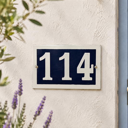 House number '114' on a blue and white sign against a light wall with plants in the background.