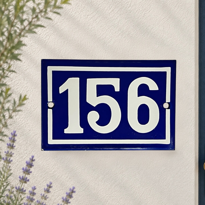 A rectangular blue house number plaque with white lettering reading '156', mounted on a textured white wall with soft leaf shadows and small plants visible at the edges.