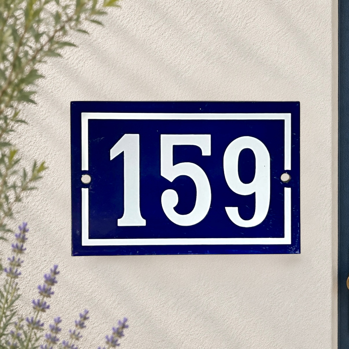 A rectangular navy blue house number plaque with white lettering reading '159', mounted on a textured off-white wall with some greenery and lavender visible at the edges.