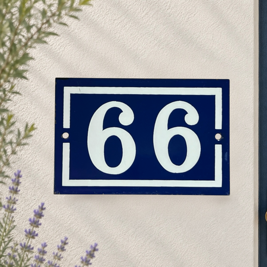A rectangular navy blue house number sign with white numerals '66' and white border details, mounted on a textured off-white wall with soft leaf shadows and small plants visible at the edges.