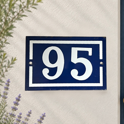 A rectangular blue house number plaque 95 with white lettering and border, mounted on a textured off-white wall with some greenery and lavender visible at the edges.
