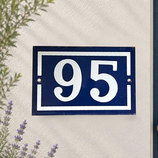 A rectangular blue house number plaque 95 with white lettering and border, mounted on a textured off-white wall with some greenery and lavender visible at the edges.