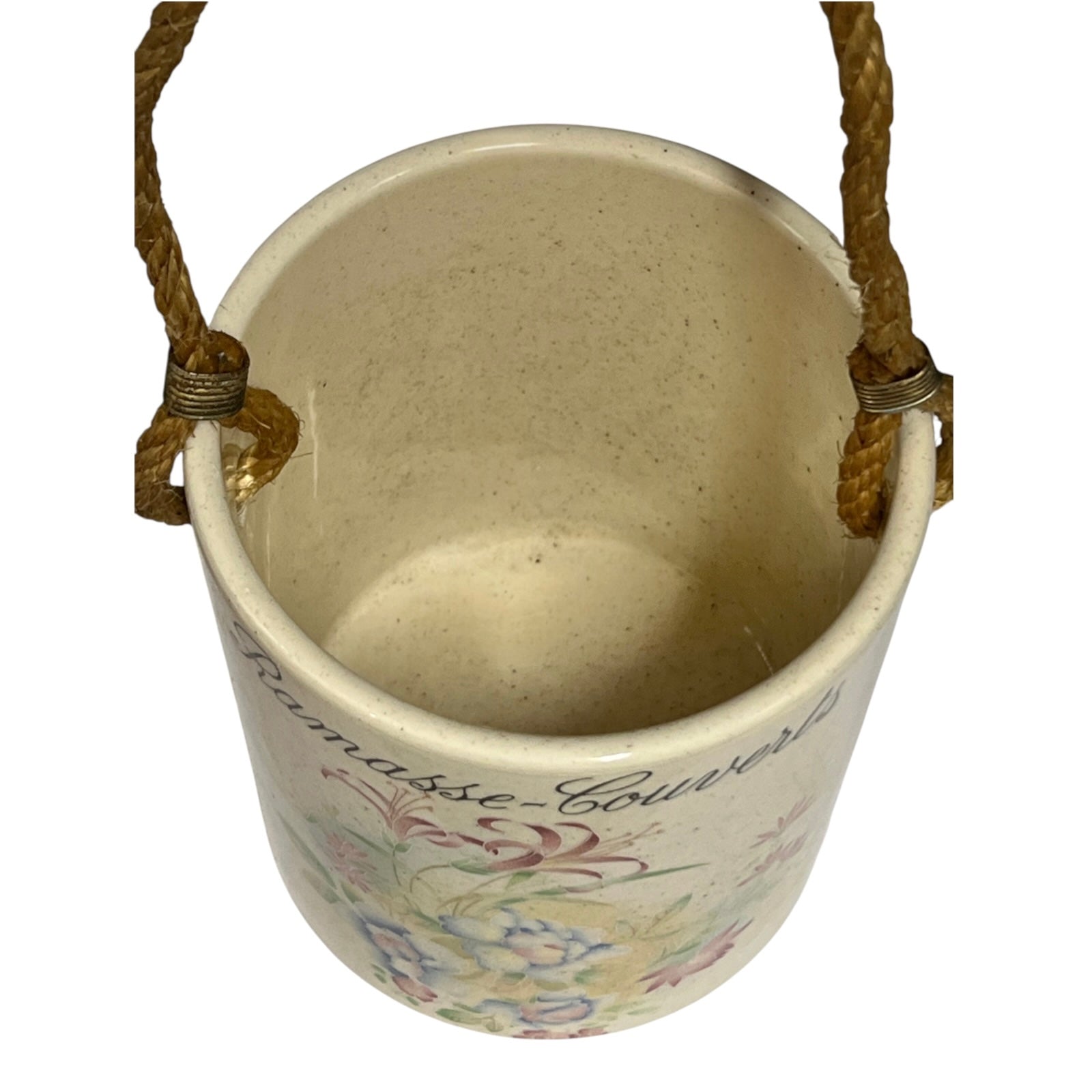 A vintage-style ceramic pot with a cream-colored glazed exterior, featuring a delicate multicolor floral pattern and the French script 'Ramasse Couvert' printed on the side. It is equipped with two braided brown rope handles secured by brass hardware, shown against a plain white background.
