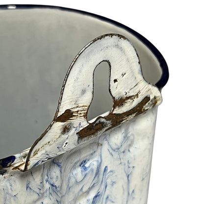 Close-up of a vintage enamel canister or cup with a white and blue marbled pattern, a dark blue rim, and a heavily distressed, chipped metal handle showing brown rusted edges.