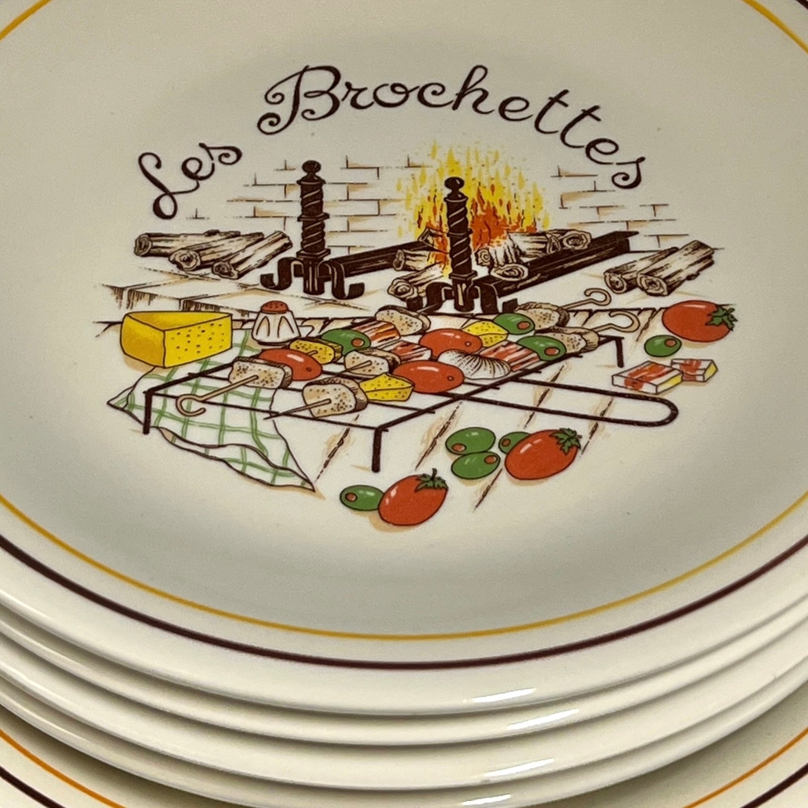 Decorative plate with 'Les Brochettes' design featuring a grill and skewers over a fire.