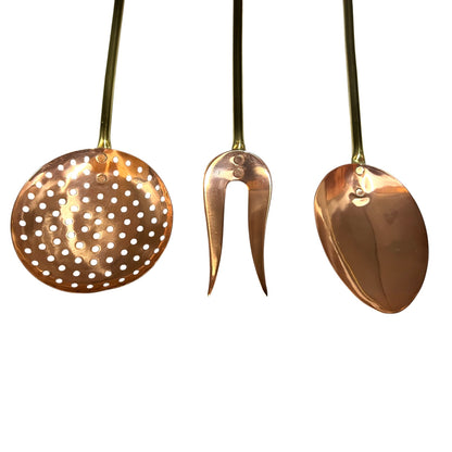 Three brass kitchen utensils with long handles, including a round perforated skimmer, a two-pronged fork, and a spoon, shown against a plain white background.