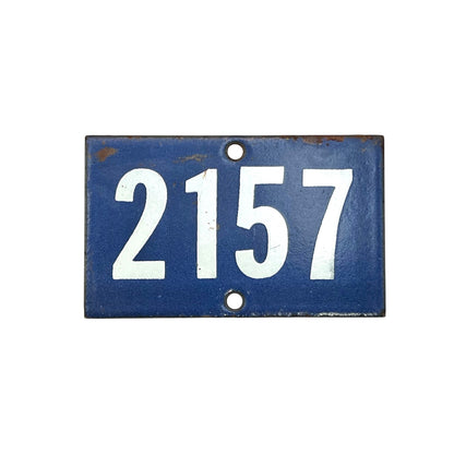 A rectangular vintage-style blue metal house number plaque with the white numbers 2157 and two pre-drilled mounting holes, showing signs of rust and age.