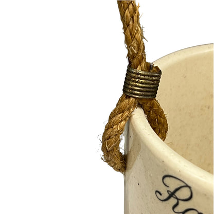 Close-up of a cream-coloured ceramic planter with a thick natural jute rope handle wrapped in a textured bronze metal band, shown against a plain white background.