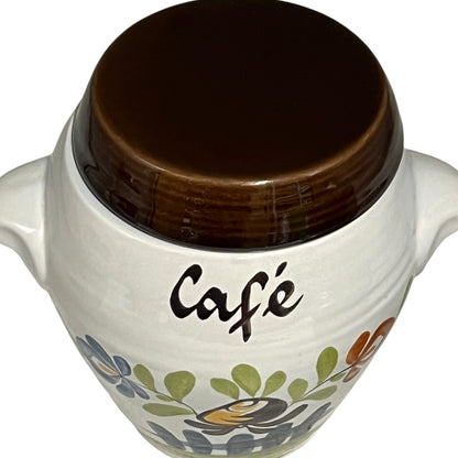 A white ceramic coffee cup or small canister with a dark brown lid, featuring the word 'Café' in black script and a floral pattern near the bottom.