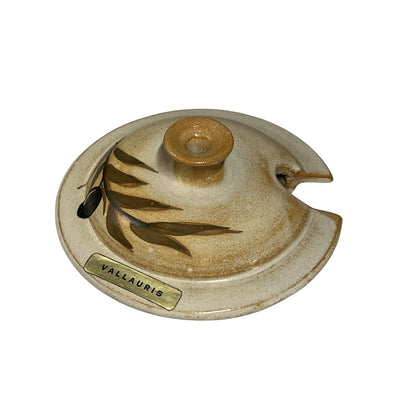 A ceramic  lid with a textured beige and brown glaze, featuring a leaf motif and a brass-colored metal brand plate reading 'VALLAURIS'.