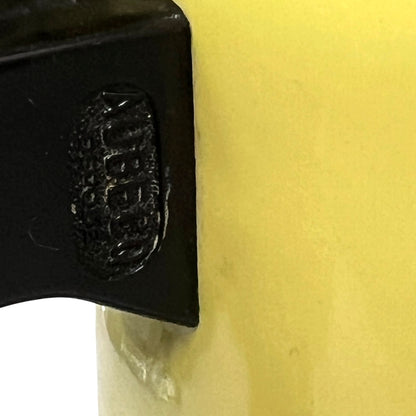 Close-up of a yellow cylindrical container with a black lid or handle attachment, showing a debossed circular logo on the black section.
