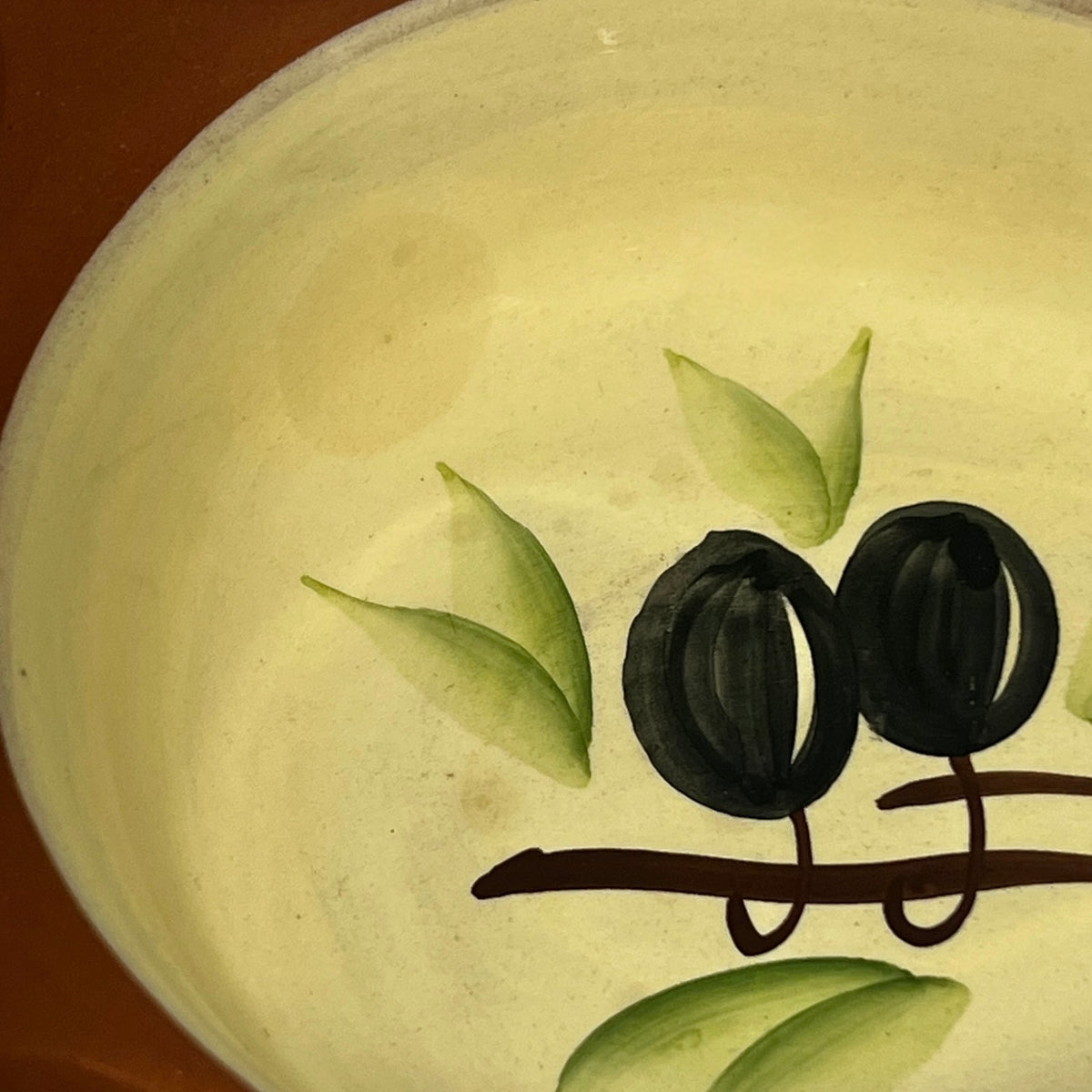 Close-up of a ceramic bowl or dish with a hand-painted design of two black olives on a brown branch with green leaves, set against a warm cream-colored glaze.