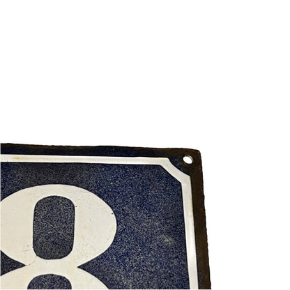 A close-up corner of a square vintage-style sign with a large white numeral 8 on a dark navy blue background, bordered by a white line and a dark edge, against a plain white background.