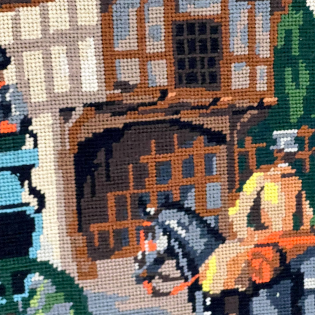 A close-up of a needlepoint or cross-stitch pattern depicting a historic building facade with timber framing, a horse, and a rider in warm earthy tones.