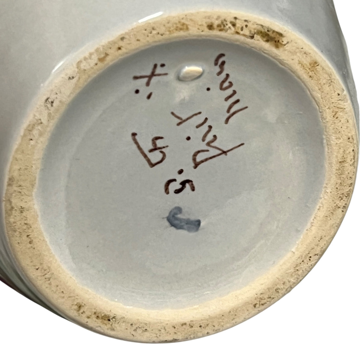Close-up of the bottom interior of a ceramic vessel showing a light gray glazed base with handwritten markings in brown and blue, plus a rough beige clay rim.