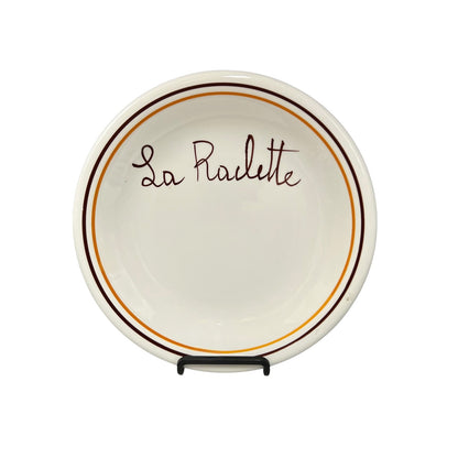 A white ceramic La Raclette plate with a dark brown and orange rim, displayed on a small black stand against a white background.