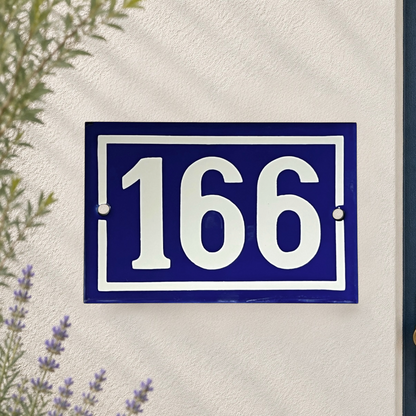 A rectangular blue house number plaque with white numerals '166' and a white border, mounted on a textured off-white wall with soft leaf shadows and small plants visible at the edges.