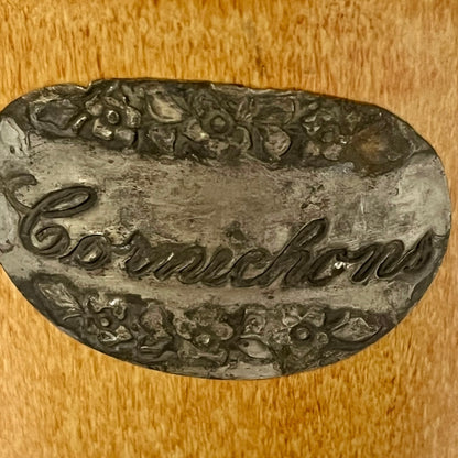 A vintage oval metal sign or plaque with the word 'Cornichons' in cursive script, decorated with embossed floral patterns at the top and bottom, shown on a wooden surface.