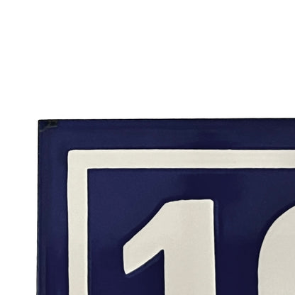 Close-up of a dark blue sign or plaque with a thick white border and part of a white numeral visible, photographed against a white background.