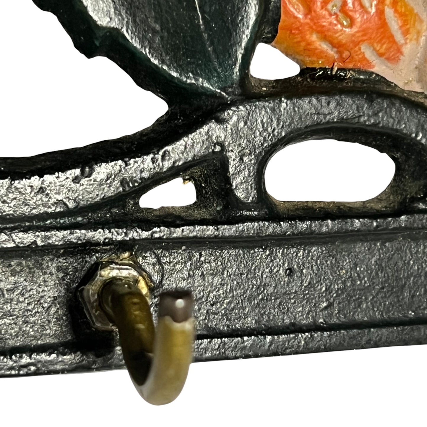 Close-up of a decorative black cast-iron piece with a small brass hook and part of an orange floral detail on a white background.