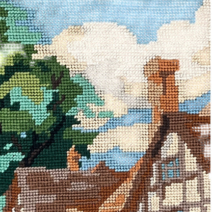 A close-up of a needlepoint canvas depicting a scenic landscape with a brown cottage, green trees, blue sky, and white clouds.