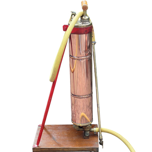 French vintage copper garden sprayer with a yellow hose and nozzle, standing upright on a wooden mount with a red-painted metal bracket. A wooden handle for pumping sits at the top, making it ideal for a vintage garden, all set against a white background.