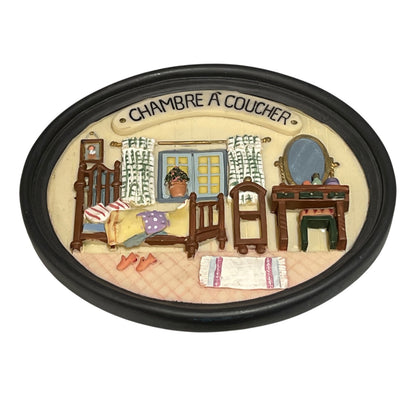 An oval decorative plaque or keepsake featuring a painted bedroom scene with the French text 'CHAMBRE A COUCHER' at the top. The illustration shows a crib, changing table, dresser, mirror, window, and small rug on a cream background, all enclosed in a black oval border.