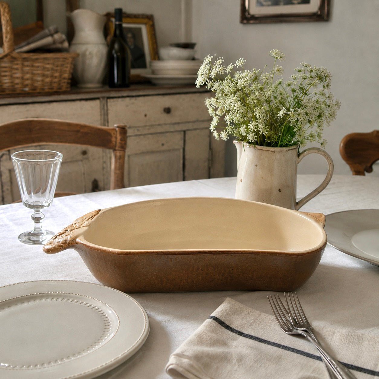 A beige ceramic 2-litre casserole dish shaped like a rabbit, featuring a brown exterior, perfect for serving game dishes or adding charm to a farmhouse-style country kitchen, displayed on a farmhouse kitchen table beside a vase of flowers
