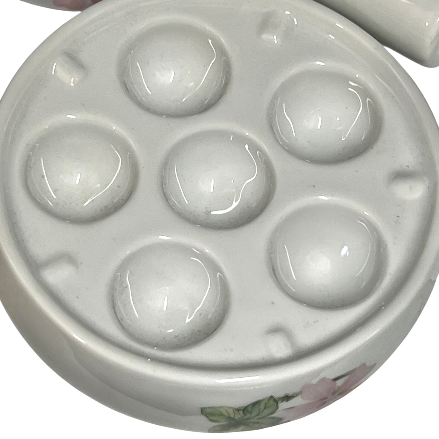 A close-up of a glossy white ceramic six-dish round palette, showing raised circular wells and a small floral decoration near the bottom edge.