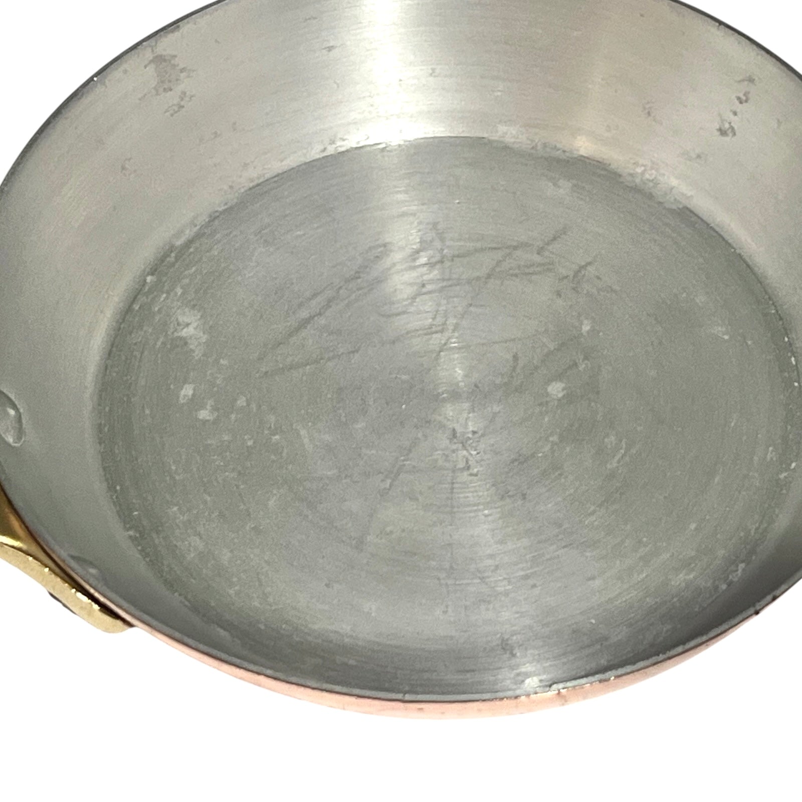 A close-up view of the inside of a copper or copper-lined saucepan with a brushed silver metal base and a small brass handle visible on the left, set against a plain white background.