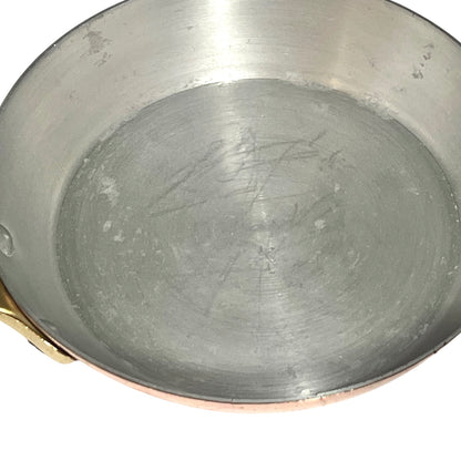 A close-up view of the inside of a copper or copper-lined saucepan with a brushed silver metal base and a small brass handle visible on the left, set against a plain white background.