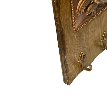 Close-up of a decorative wall-mounted wooden board with carved relief details and small hanging hooks, shown against a plain white background.
