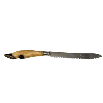 A long serrated steak knife with a polished silver steel blade and a handle made of natural tan and black animal fur, shown horizontally against a plain white background.