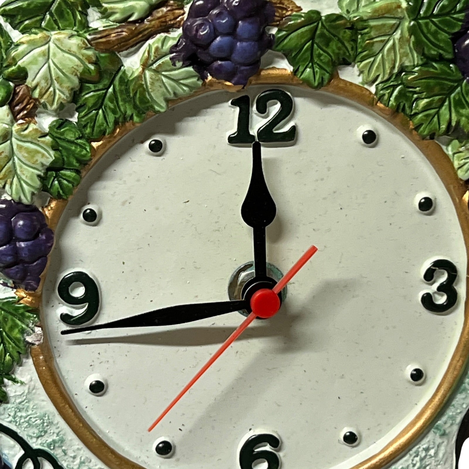 Close-up of a decorative wall clock face with black numerals, black hour and minute hands, a red second hand, and a gold-toned rim. Grapevine and grape motifs decorate the surrounding border.