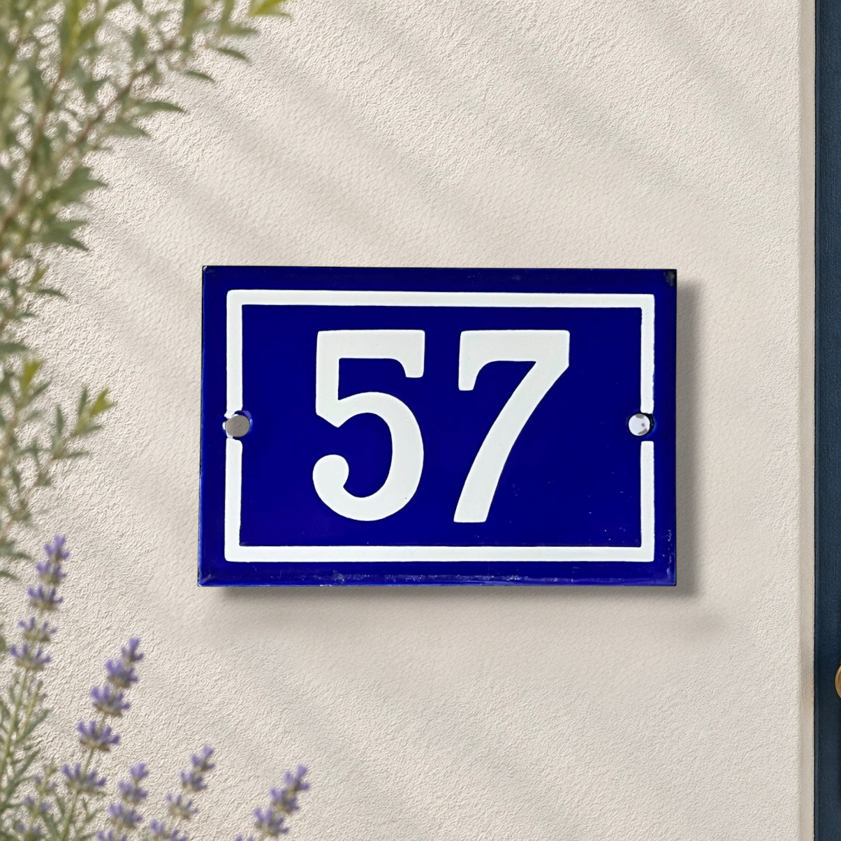 A rectangular blue house number plaque with white numerals '57' mounted on a textured off-white wall, with soft green leaves and lavender flowers framing the scene.