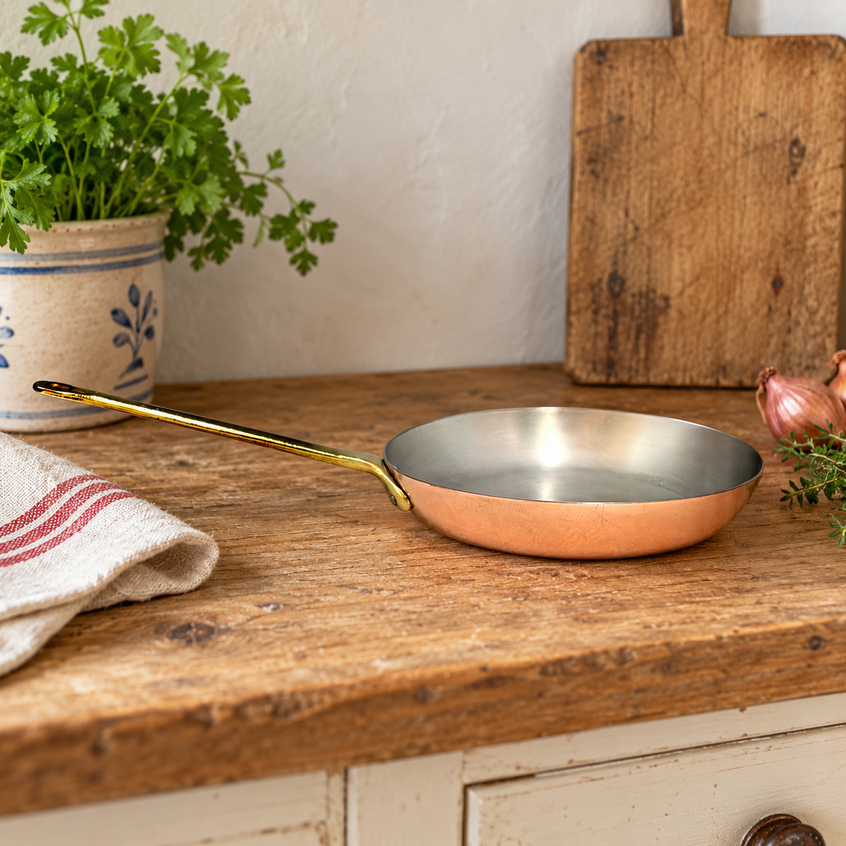 A copper frying pan with a polished gold handle and silver interior, resting on a rustic wooden countertop with kitchen props including a potted herb, a cloth towel, and a wooden cutting board.