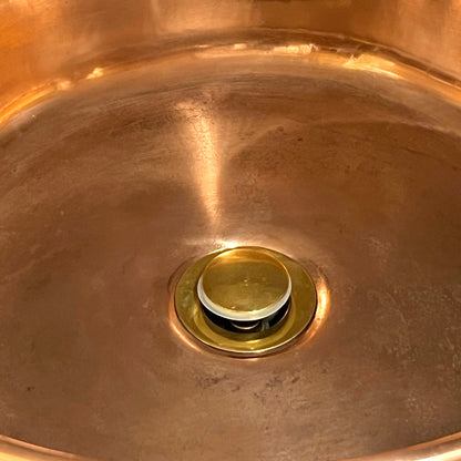 A close-up view of a round brass kitchen sink drain set into a copper sink basin.