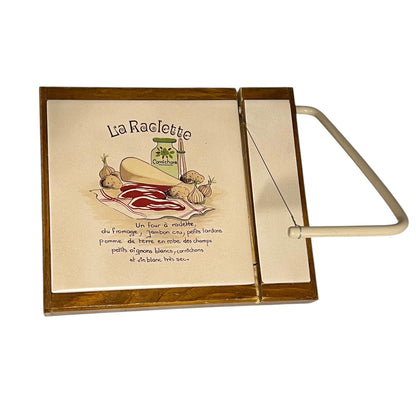 A wooden La Raclette cheese board set on a white background, featuring a square wooden box with a cream-coloured interior printed with French cheese ingredients and illustrations, alongside a matching wooden handle and wire