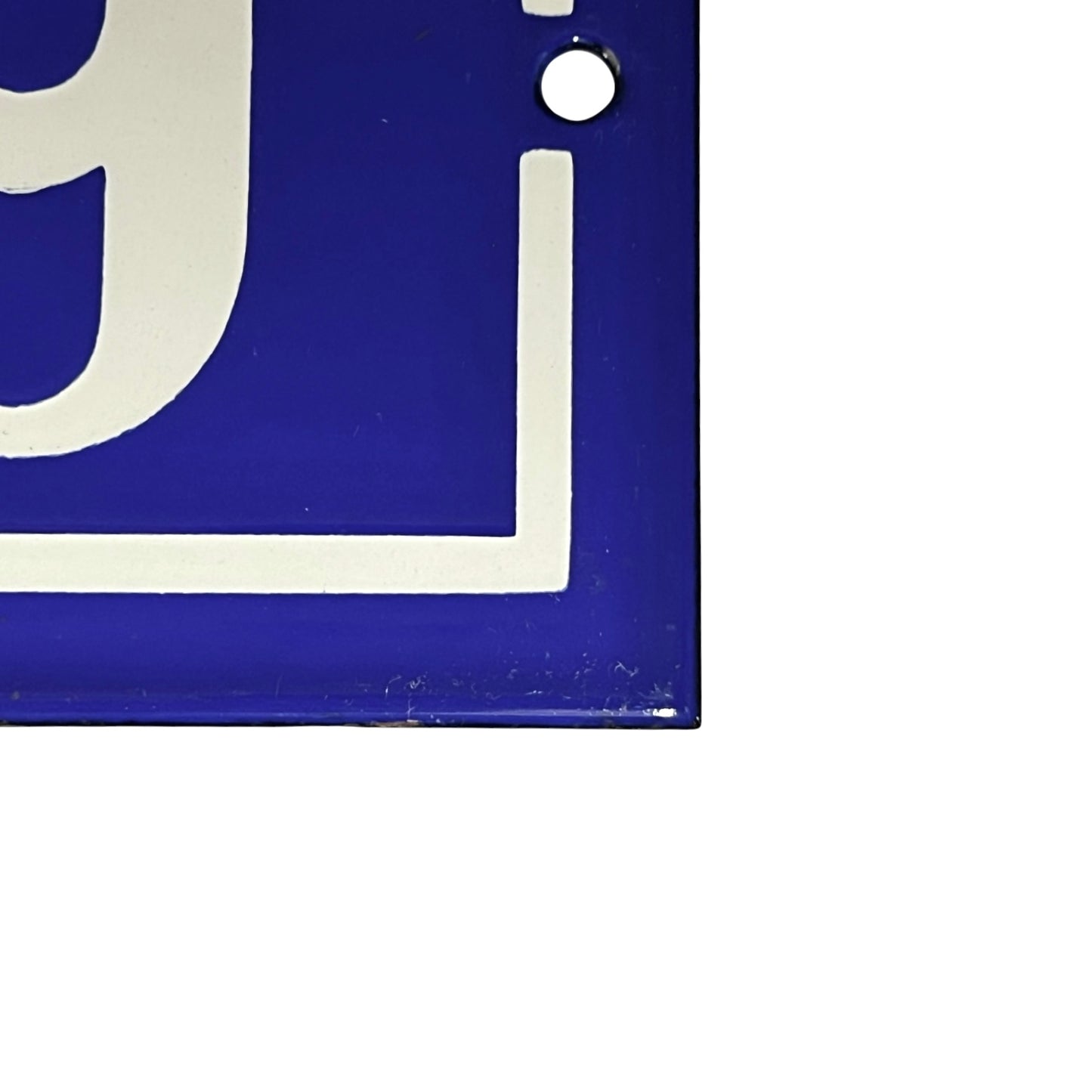 Close-up of a blue square sign or plaque with a white border and partial white lettering, showing a small mounting hole near the top right, on a white background.