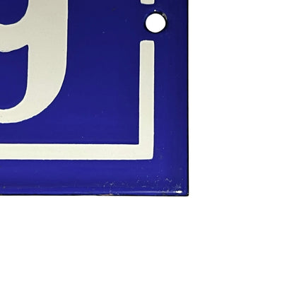 Close-up of a blue square sign or plaque with a white border and partial white lettering, showing a small mounting hole near the top right, on a white background.