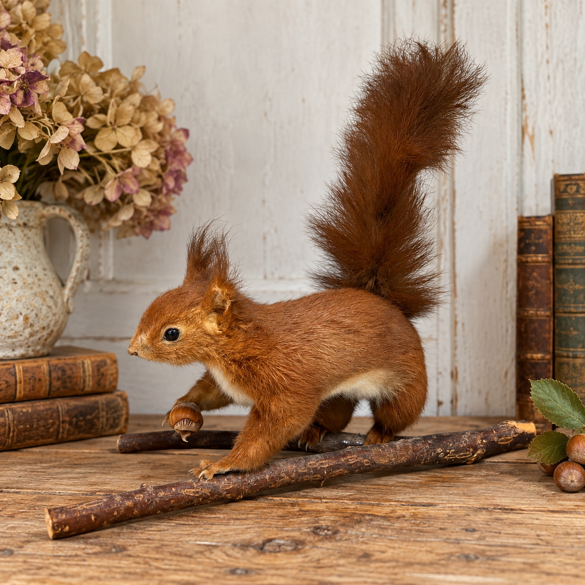 A taxidermy red squirrel taxidermy decoration standing on a rustic wooden branch on a wooden table, with vintage books, a ceramic vase of dried flowers, and acorns in the background.
