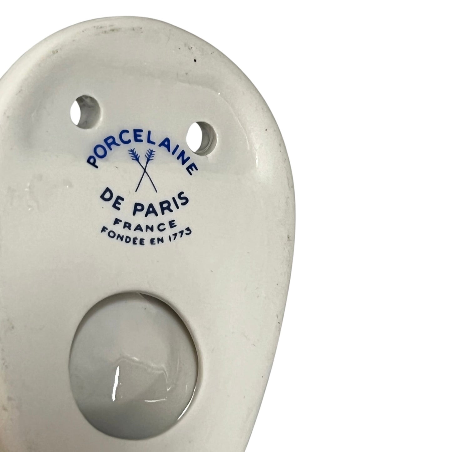 Close-up of the back of a white porcelain toilet paper holder with blue printed text reading 'PORCELaine DE PARIS FRANCE FONDÉE EN 1775' on a white background.
