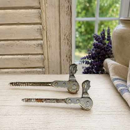 Pair of vintage-style metal hinges on a wooden surface with a window and lavender in the background.