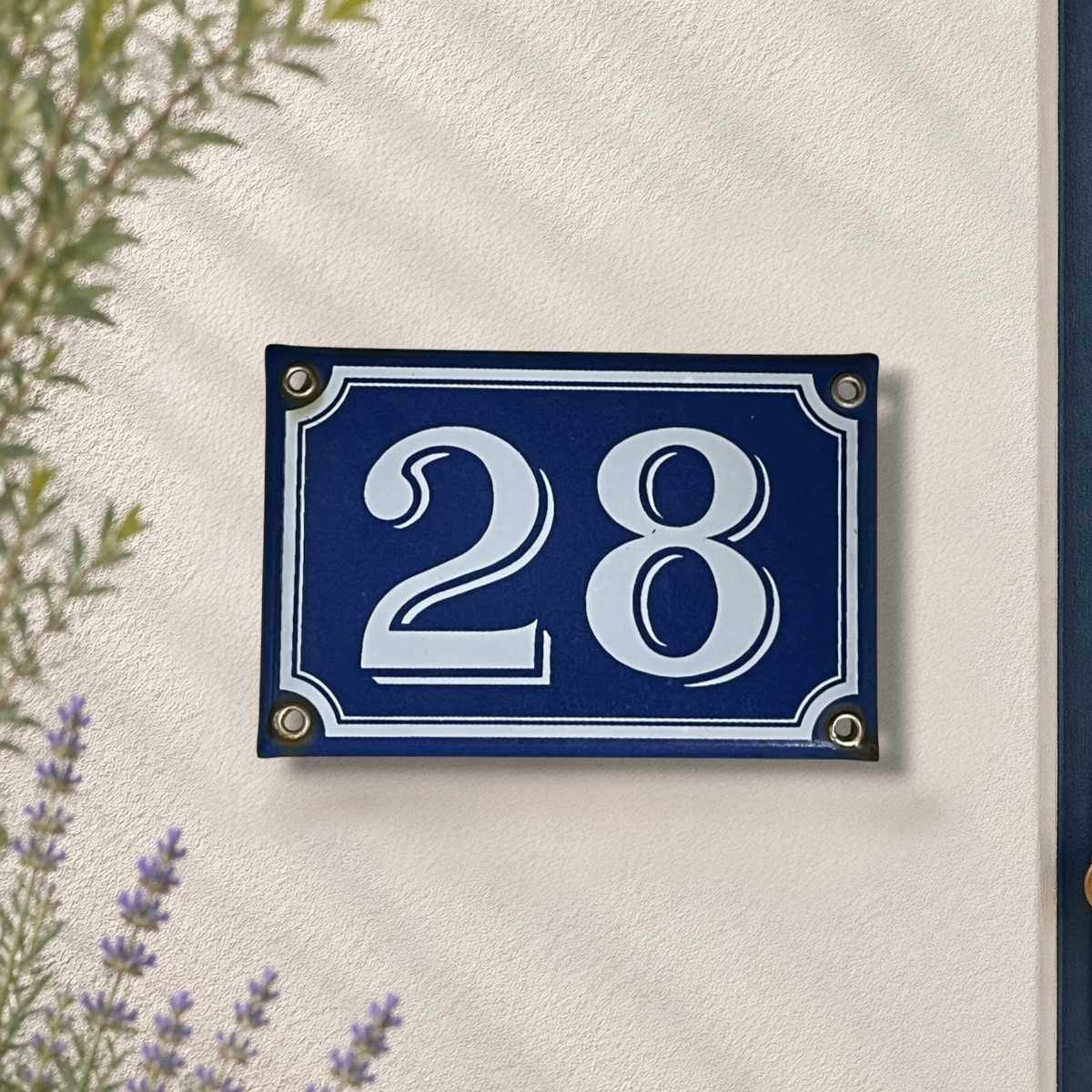 A rectangular blue house number plaque with white numerals '28' and white border details, mounted on a textured off-white wall with soft leaf shadows. Small decorative plants are visible at the edges.