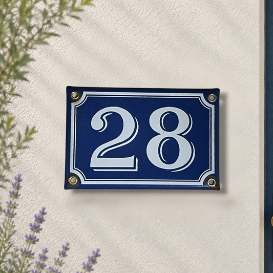A rectangular blue house number plaque with white numerals '28' and white border details, mounted on a textured off-white wall with soft leaf shadows. Small decorative plants are visible at the edges.