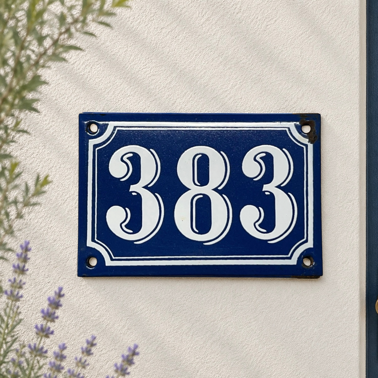A rectangular blue house number plaque with white decorative numerals and border, mounted on a textured off-white wall with some greenery and lavender visible at the edges.