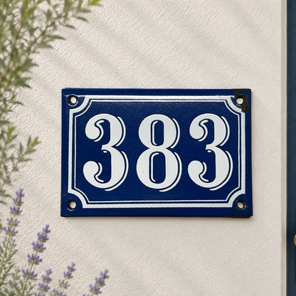 A rectangular blue house number plaque with white decorative numerals and border, mounted on a textured off-white wall with some greenery and lavender visible at the edges.