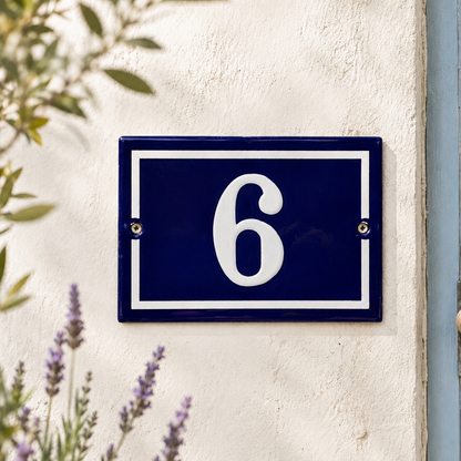 Blue enamel French-style house number plaque displaying the number 6 in white, mounted on a cream rendered exterior wall with visible fixing screws. Soft-focus lavender flowers and leafy branches frame the sign in natural daylight