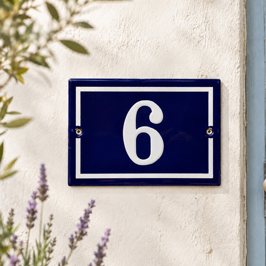 Blue enamel French-style house number plaque displaying the number 6 in white, mounted on a cream rendered exterior wall with visible fixing screws. Soft-focus lavender flowers and leafy branches frame the sign in natural daylight