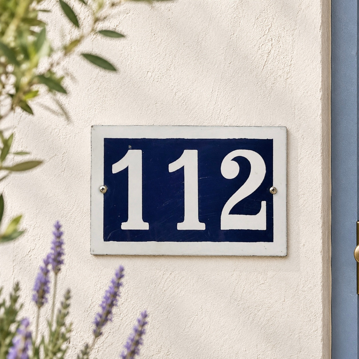 House number '112' on a blue and white sign mounted on a wall with plants in the background.